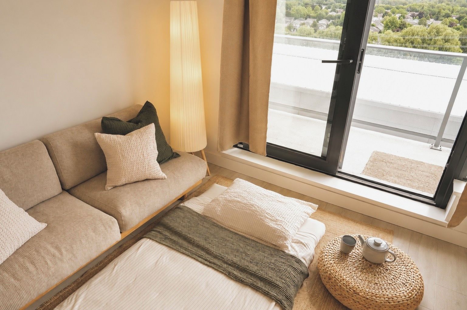 Een hotelkamer met een bank, een raam met uitzicht op groen, een lamp en een klein rond tafeltje met een theepot.