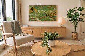 Ein modernes Wohnzimmer mit einem beigen Sofa, einem runden Holztisch, einer Topfpflanze, einem Gemälde an der Wand und einer Lampe.