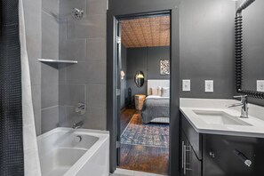 A modern bathroom with a bathtub, a shower, and a sink. There is a wooden floor and a bed visible through an open door. - Luxury Nashville Urban Cowboy Rooftop | Sleeps 10 (Nashville)