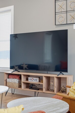 Ein modernes Wohnzimmer mit einem großen Flachbildfernseher auf einem Holz-TV-Schrank, einem kleinen runden Tisch und einer Couch mit Kissen.