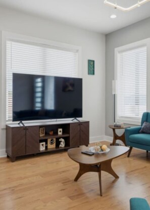 A modern living room with a flat-screen TV on a wooden cabinet, a round coffee table, and a teal armchair. - WeHo 8BR Retreat | Two Homes, Room for 24 (Nashville)