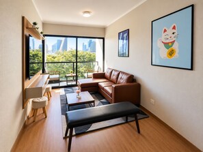 Ein modernes Wohnzimmer mit einem braunen Ledersofa, einer schwarzen Bank und einem Couchtisch. Ein großes Fenster bietet Blick auf Gebäude und Bäume.