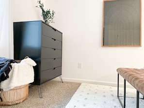 A bedroom with a dark dresser, a bench, a plant, and a framed artwork on the wall.