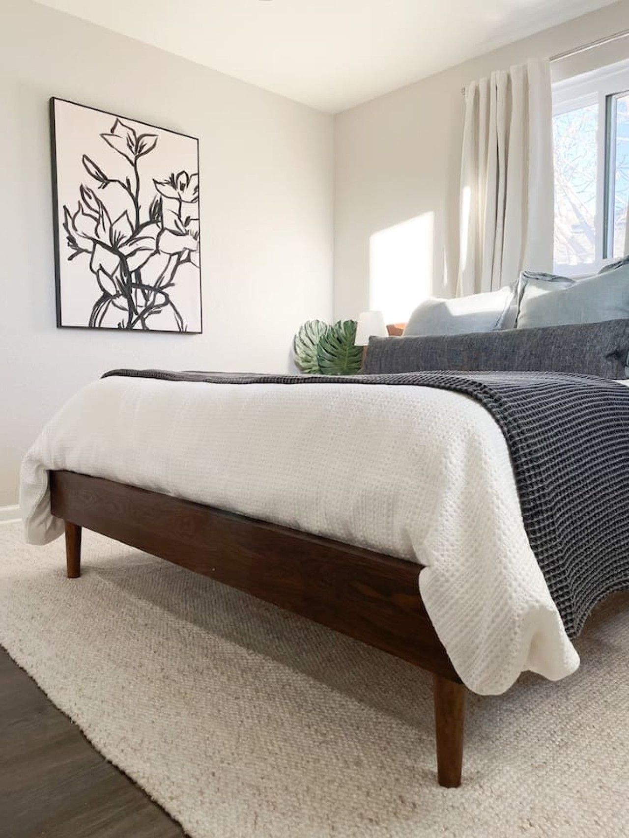Une chambre à coucher avec un lit, une grande fenêtre avec des rideaux, une œuvre d’art accrochée au mur et une plante.