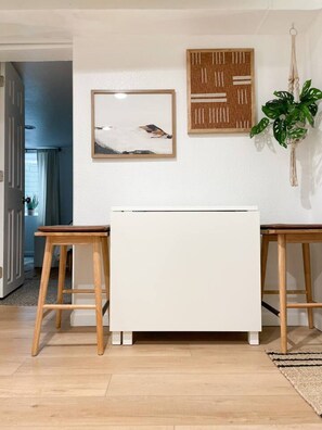 A room with a white cabinet, two wooden stools, a framed abstract painting, a macrame plant hanger, and a door leading to another room.
