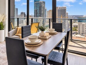 Un coin repas avec une table dressée pour deux personnes, des chaises, un vase rempli de fleurs et une vue sur le paysage urbain grâce à de grandes fenêtres.