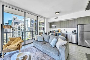 A modern apartment with a grey sofa, a wooden chair, a kitchen with stainless steel appliances, and a large window offering a city view. - High Rise Ocean View Downtown Miami (Miami)