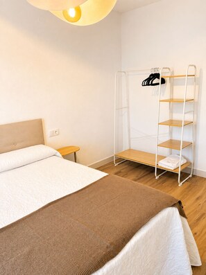 A bedroom with a bed, a small bedside table, a hanging clothes rack, and a metal shelf unit. - ALBACETELOFT Modern 2-bedroom apartment | Wi-Fi | Ideal for Work and Family (Albacete)