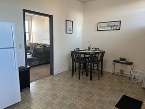 A compact kitchen with a refrigerator, a dining table with chairs, a small trash bin, and a laundry basket. - The Downtown Hideaway – Stylish 1BR Comfort (Greensboro)