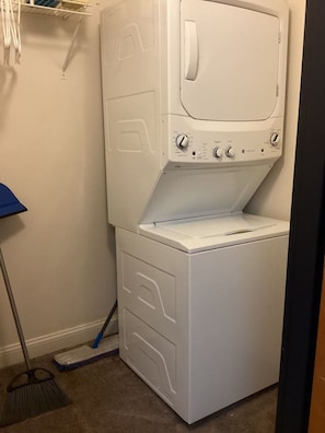 A compact, white laundry room with a built-in washer and dryer, a broom, and a mop on the floor. - Downtown Lounge Getaway with Two Living Spaces (Greensboro)