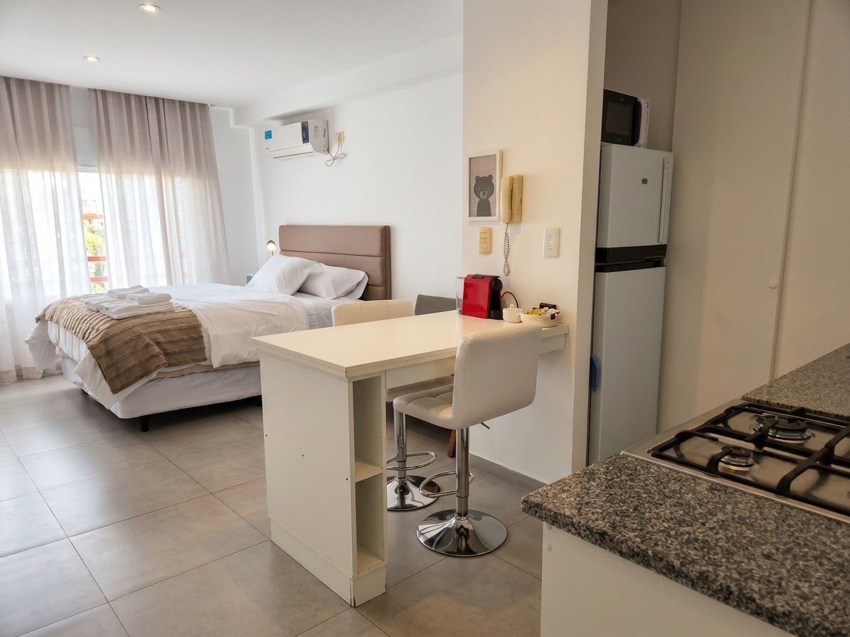 A modern hotel room with a bed, a small kitchenette, a dining area, and a bathroom.