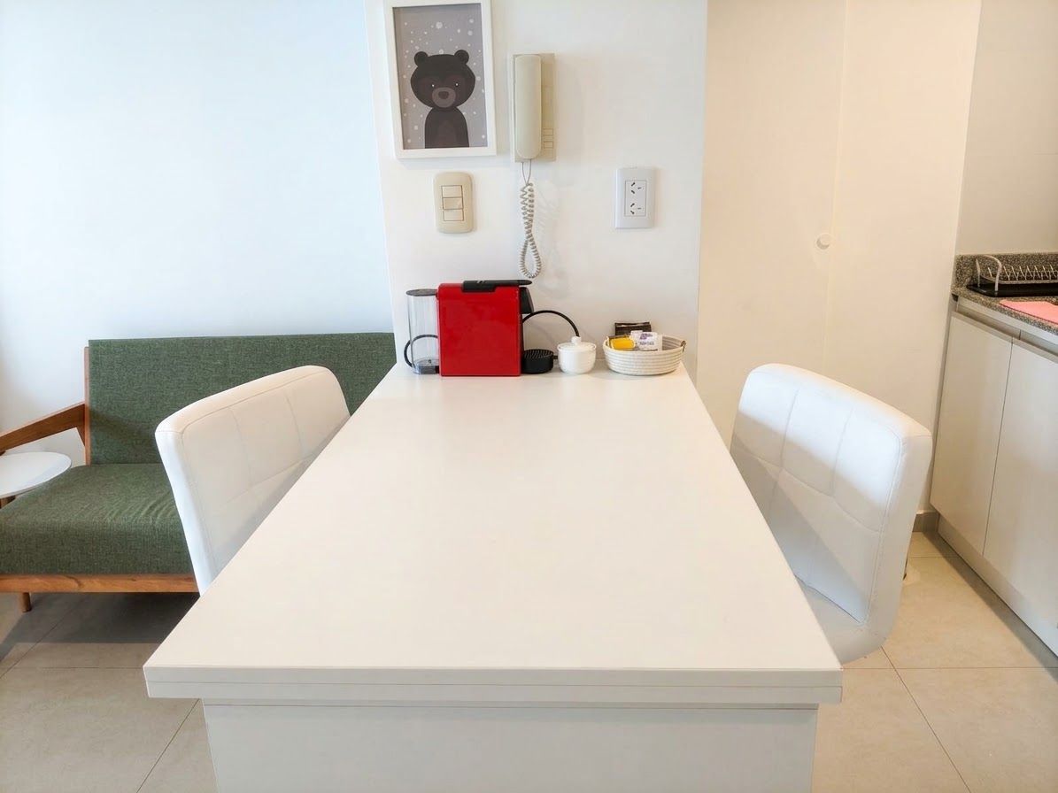 Une pièce moderne et minimaliste, avec une table blanche, deux chaises blanches, un canapé vert et un sac rouge posé sur la table.