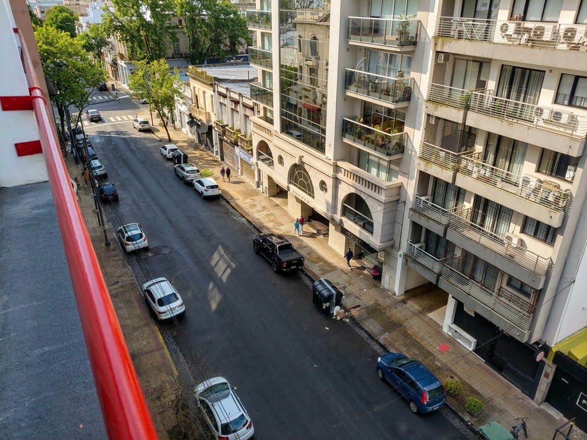 Un immeuble à plusieurs étages doté de balcons, une rue bordée de voitures garées et des piétons.