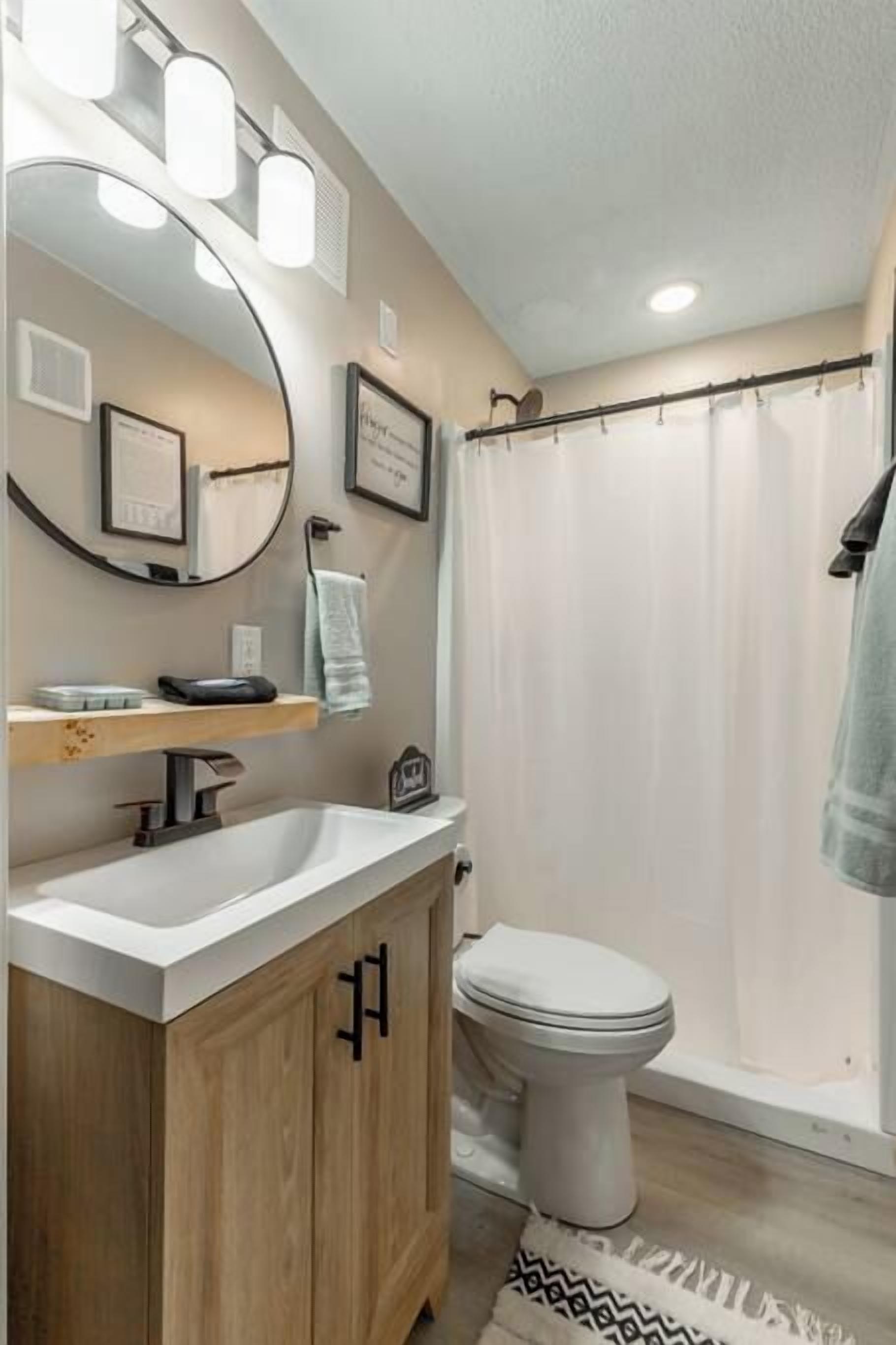 A bathroom with a wooden vanity, a white toilet, a shower curtain, and a mirror with lights.