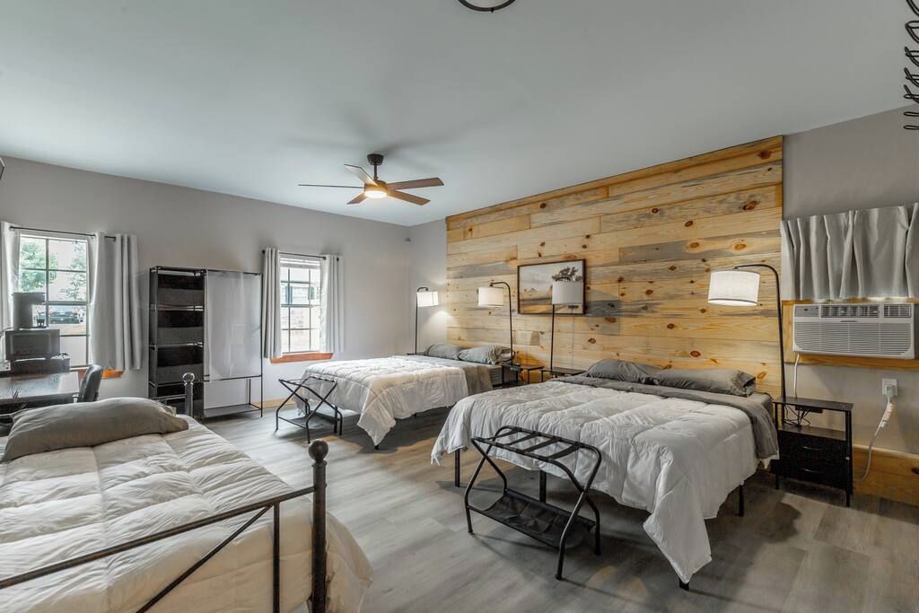 A room with two beds, a ceiling fan, a wooden wall panel, and a desk with a chair.
