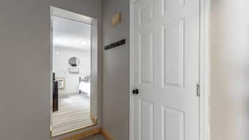 Un couloir avec une porte blanche, une petite étagère avec quelques objets, et une vue sur une pièce où l’on aperçoit un miroir et une chaise.