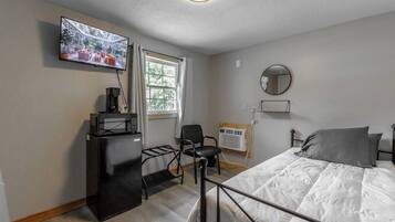 Une chambre à coucher avec un lit, un téléviseur fixé au mur, un bureau avec un ordinateur et une fenêtre avec des rideaux.
