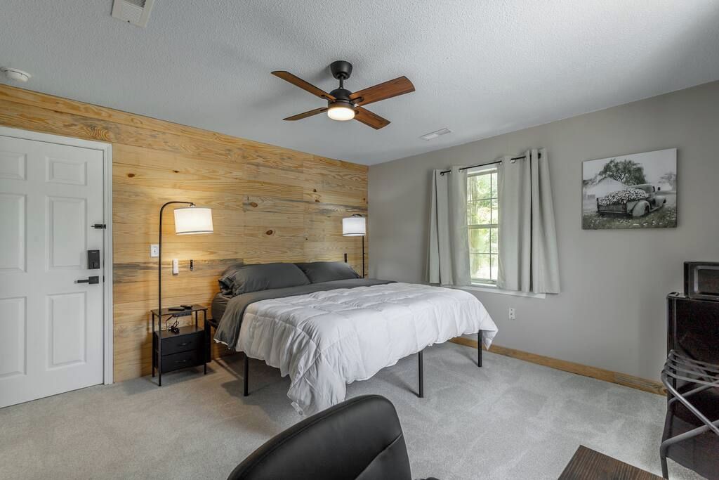 A bedroom with a bed, a ceiling fan, a nightstand, a desk, and a window with curtains.