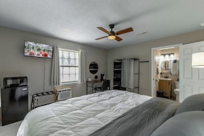 Un dormitorio con cama, ventilador de techo, televisor, escritorio, silla, ventana y baño.