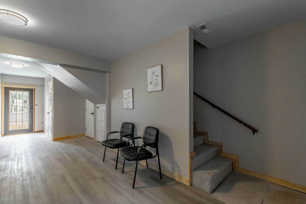 Un espacio interior con una escalera, dos sillas y una puerta que da a una zona luminosa.