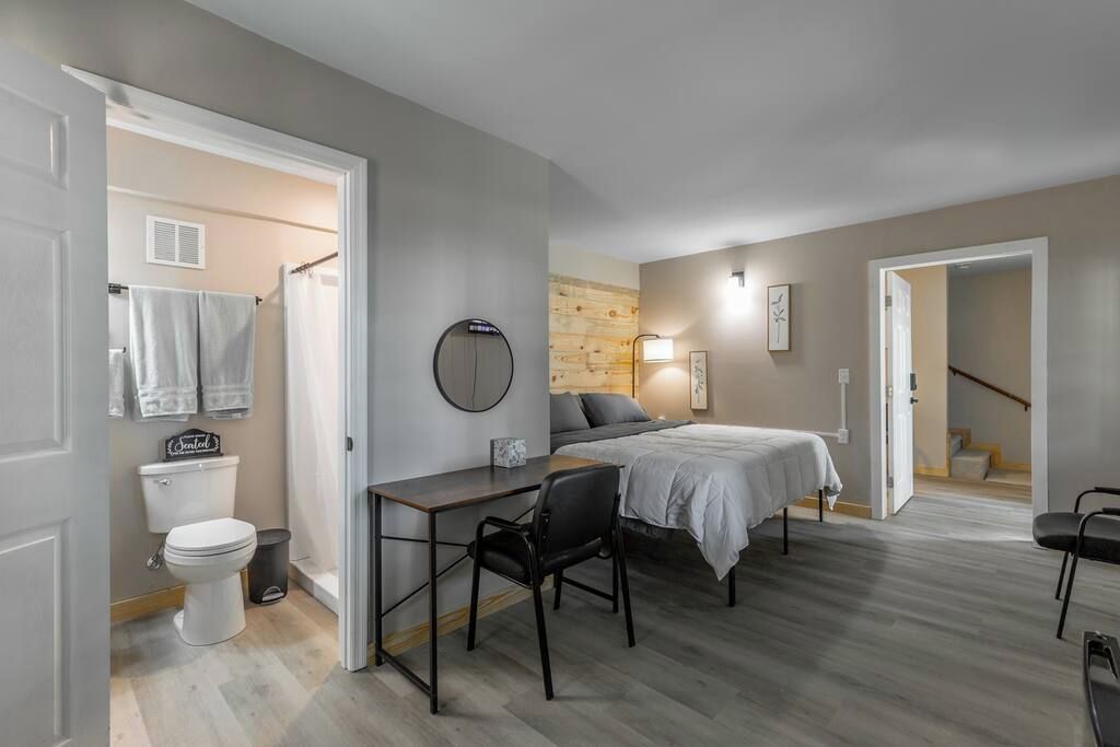 Una habitación compacta con cama, escritorio y silla. Se ve un baño con inodoro y toallero.