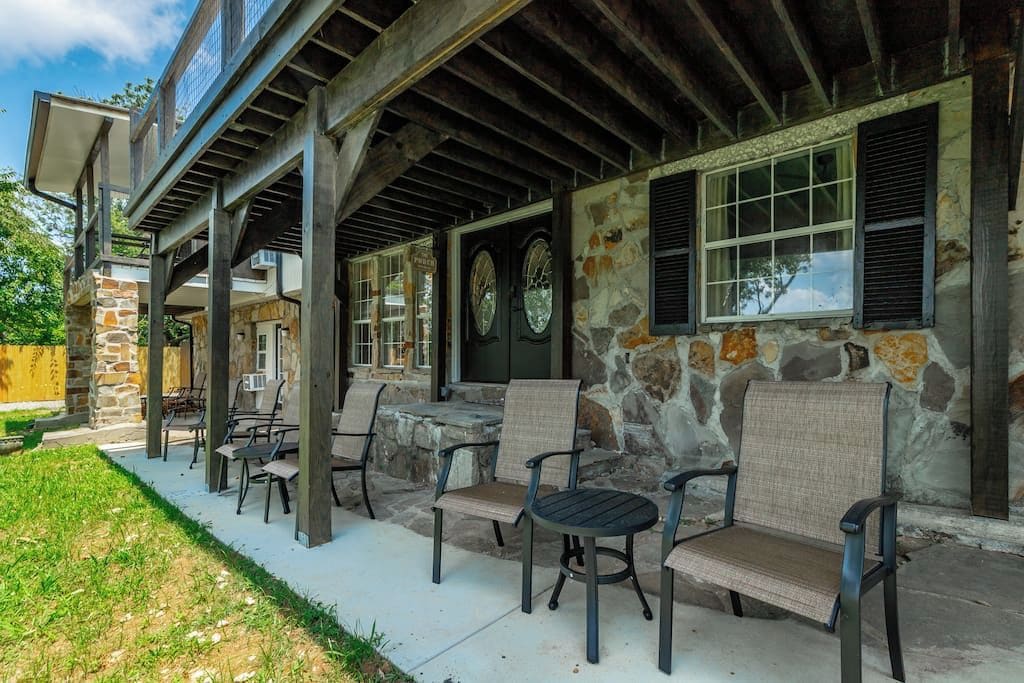 A covered outdoor seating area with stone walls and wooden beams, featuring several chairs and tables.