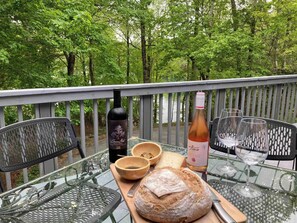 Une table en verre sur laquelle se trouvent un pain, une bouteille de vin et des verres, le tout placé sur un balcon offrant une vue sur une forêt.