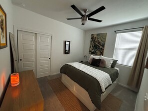 Ein Schlafzimmer mit einem Bett, einem Deckenventilator, einem Fenster mit Jalousien, einer Tür und einem gerahmten Bild an der Wand.