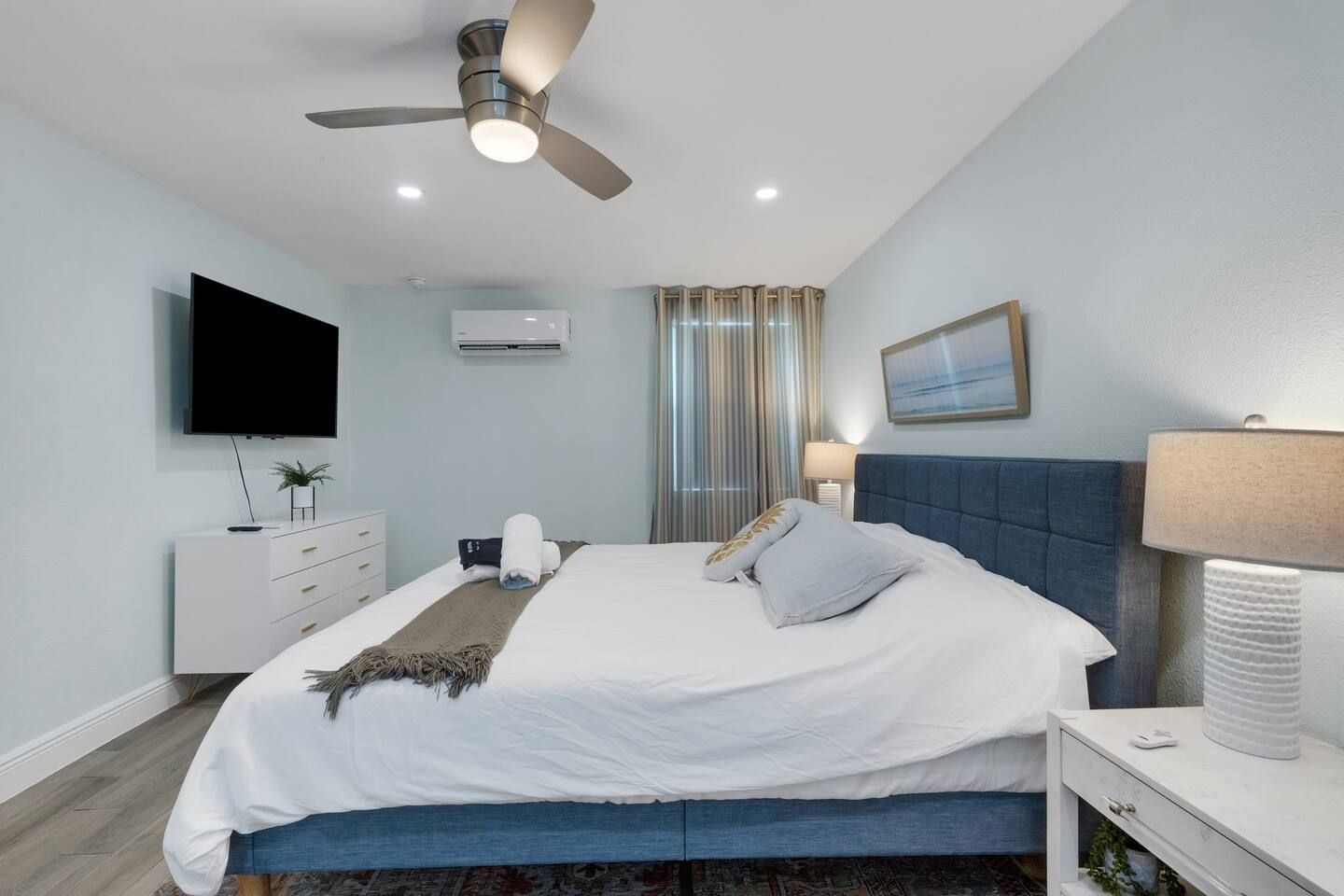 A modern hotel room with a bed, a flat-screen TV, a ceiling fan, an air conditioning unit, a bedside lamp, and a window with curtains.
