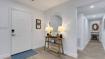 Um corredor com porta branca, mesa consola, luminárias, espelho e tapete azul.