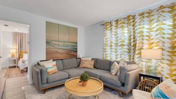 Uma sala de estar moderna com um sofá seccional cinza, uma mesa de centro de vime redonda e uma grande pintura abstrata na parede.