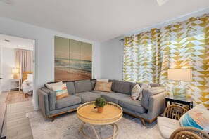 Een moderne woonkamer met een grijze hoekbank, een ronde rieten salontafel en een groot abstract schilderij aan de muur.