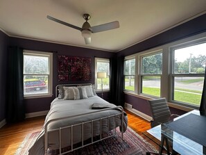 Une chambre à coucher avec un lit, un bureau et une chaise. Elle est dotée de grandes fenêtres et d’un ventilateur de plafond.