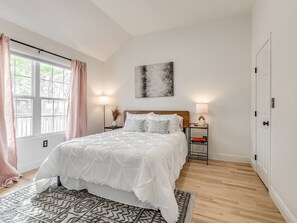 Une chambre à coucher avec un lit, des tables de chevet, une commode, une fenêtre avec des rideaux, un tableau au mur et une porte.