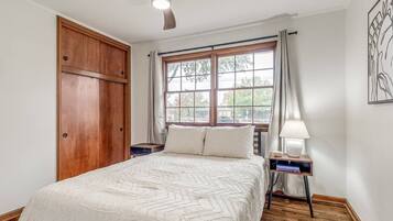Un dormitorio con cama, ventilador de techo, ventana con cortinas, mesita de noche con lámpara y armario de madera.