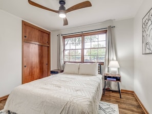Een slaapkamer met een bed, een plafondventilator, een raam met gordijnen, een nachtkastje met een lamp en een houten garderobe.
