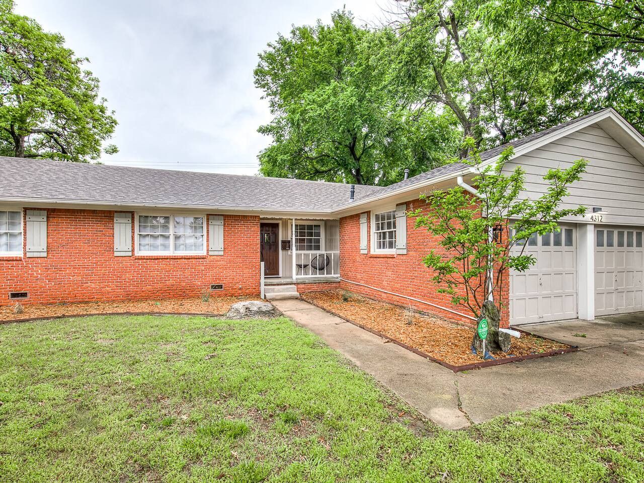 A single-story brick house with a front porch, a garage, and a small tree in the yard.