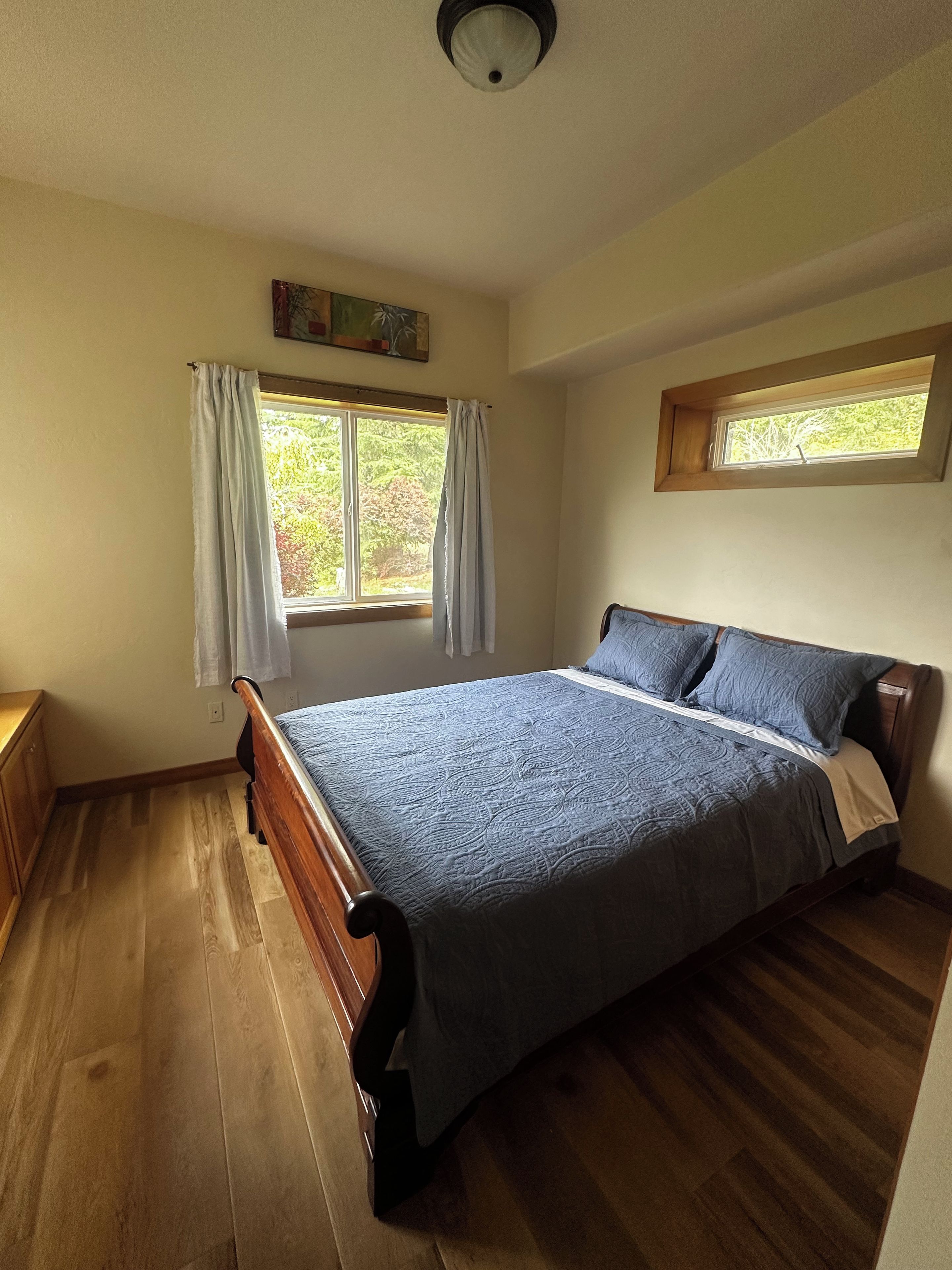 Ein Schlafzimmer mit einem hölzernen Bett, zwei Fenstern mit Vorhängen, einer Deckenleuchte und einem kleinen Oberlicht.