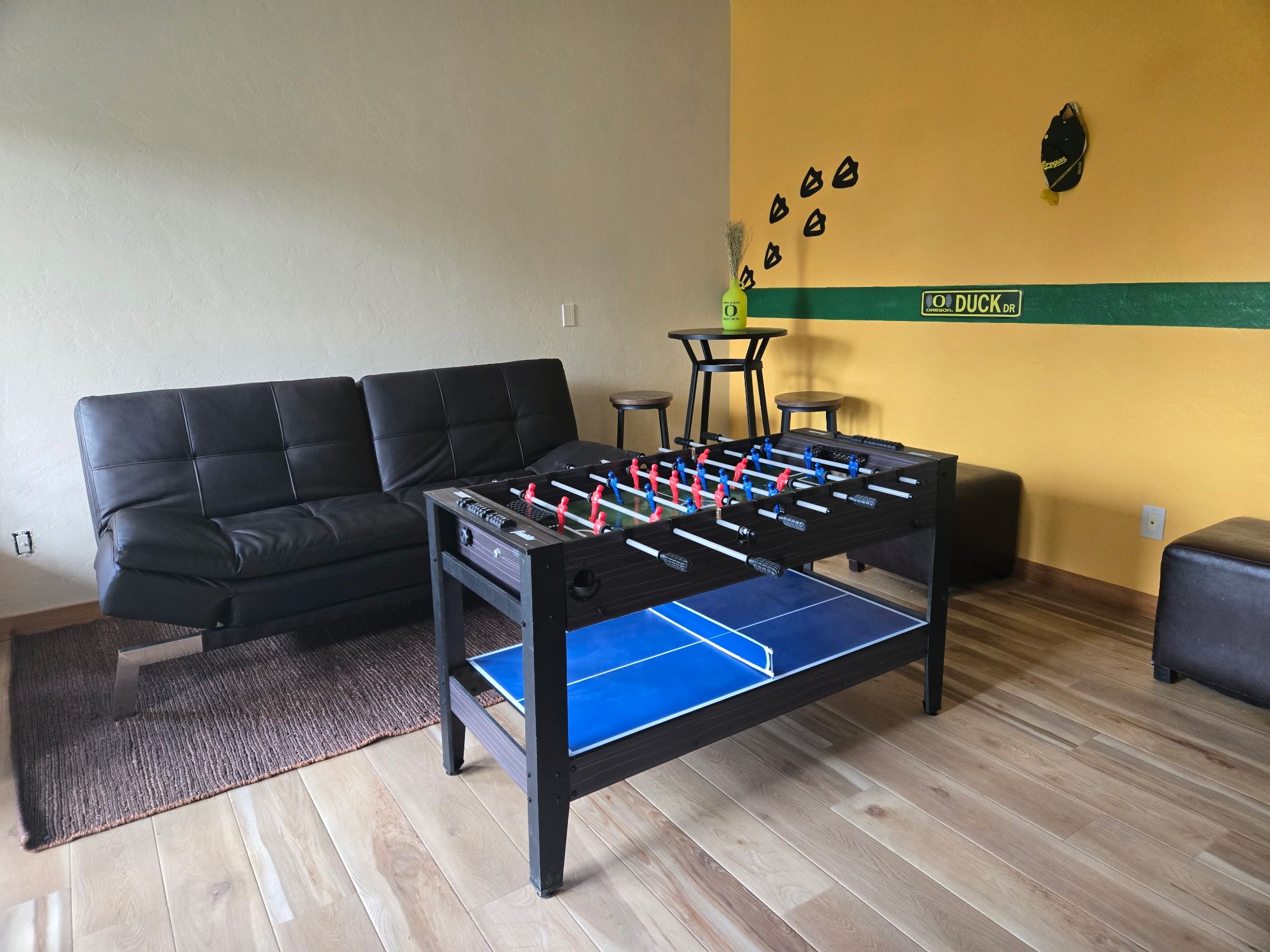 Quarto com mesa de futebol de mesa, sofá preto, pequena mesa redonda e parede com faixa verde e um logotipo.