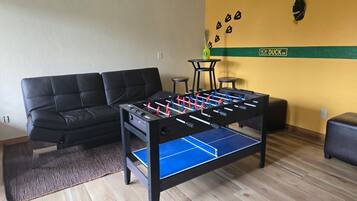 Quarto com mesa de futebol de mesa, sofá preto, pequena mesa redonda e parede com faixa verde e um logotipo.