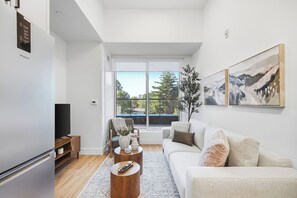 A modern living room with a flat-screen TV, a sofa, a coffee table, and a large window offering a view of trees and buildings. - Downtown 1BR | Gym + Coworking Area + Balcony (Kitchener)