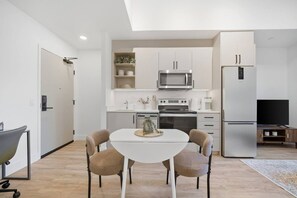 A modern kitchen with white cabinets, a microwave, stove, and a refrigerator. There is a dining table with four chairs and a small rug on the floor. - Downtown 1BR | Gym + Coworking Area + Balcony (Kitchener)