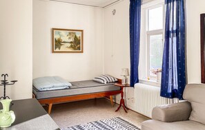 Une petite pièce bien éclairée, avec un lit, une table de chevet dotée d’une lampe, une fenêtre avec des rideaux, un radiateur et un tableau accroché au mur.
