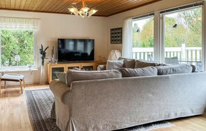A living room with a large grey sofa, a flat-screen TV on a wooden stand, a small table with a lamp, and a view of greenery through large windows. - 4 bedroom gorgeous home in örkelljunga (Örkelljunga)