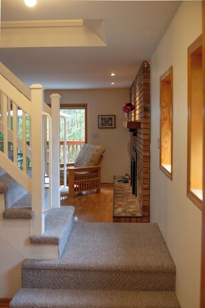 Una escalera conduce a una sala de estar con chimenea, un banco de madera y un ventanal con vistas a la vegetación.