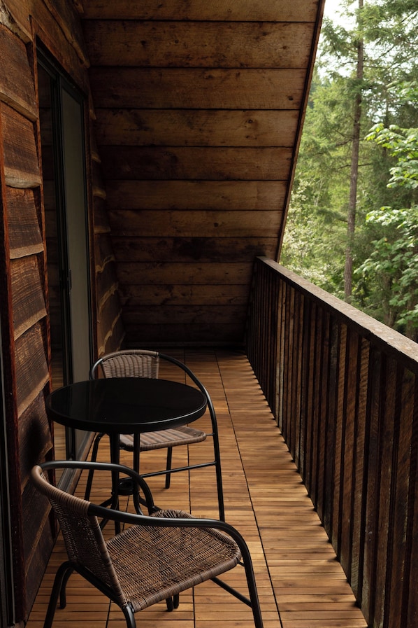 Una terraza de madera con dos sillas de mimbre y una mesa, con vistas a un área boscosa.