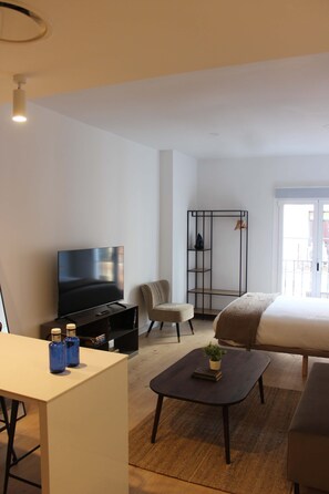 A modern hotel room with a bed, a TV, a seating area with a chair, a small table, and a bookshelf. - w* | Bright 1BR with Balcony in Central Madrid (Madrid)