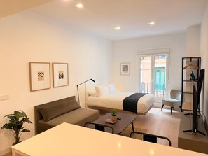 A modern hotel room with a bed, a sofa, a small table, and a desk. There are two framed pictures on the wall, a plant, and a balcony with a view. - w* | Airy 1BR w/ Balcony in Central Madrid (Madrid)
