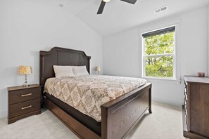 A bedroom with a wooden bed, two bedside tables with lamps, a window with a view of trees, and a ceiling fan. - Music City Home Base | Office + 98" TV | 30+ Days (Nashville)