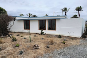 A single-story building with a flat roof, three windows with wooden frames, and a sandy ground with sparse vegetation. - Casa Frida - Beach City retreat (Ensenada)